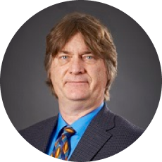 A person with long brown hair is wearing a dark suit and a colorful tie, posing against a plain gray background.