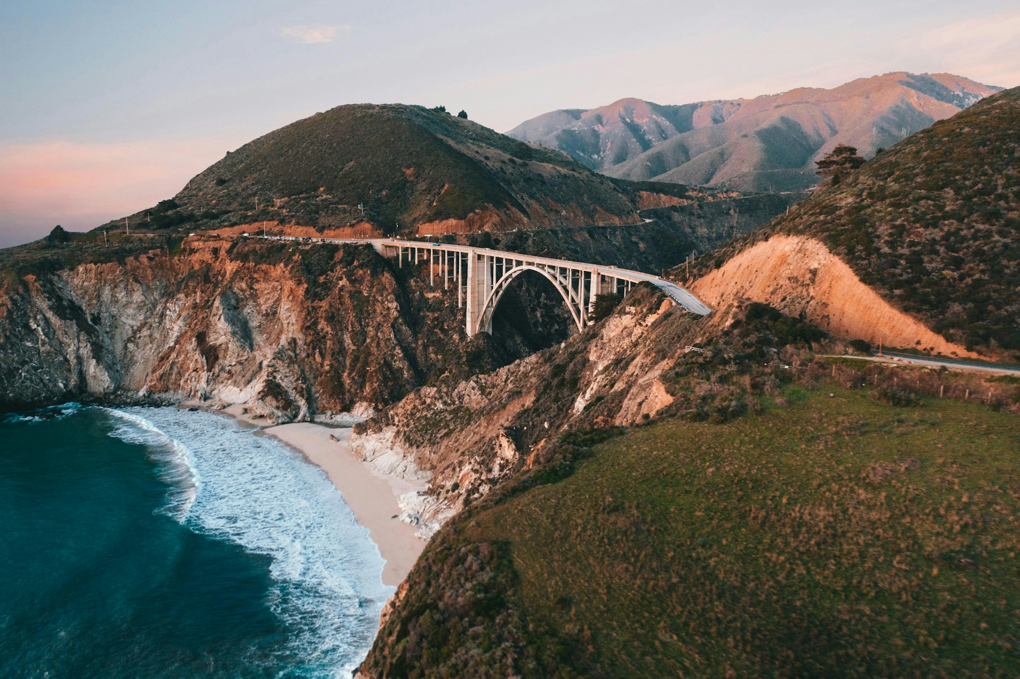 Coastal landscape with a bridge spanning rugged cliffs, overlooking a beach and ocean waves. Photo by: Spencer Davis