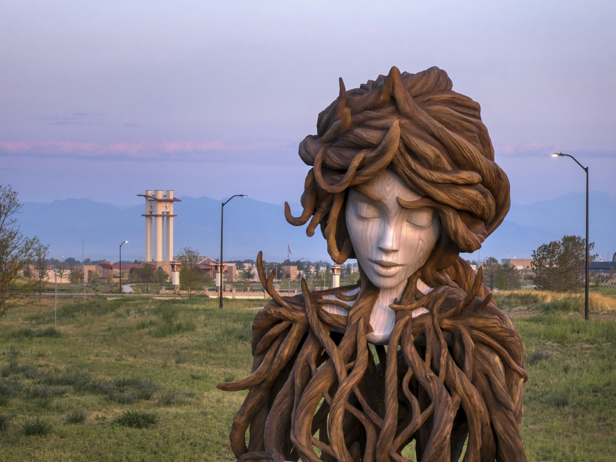 A sculpture of a woman with intricate hair design set in a grassy landscape with a building and mountains in the background.