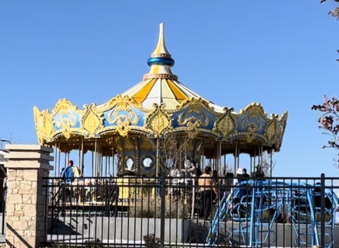 A colorful carousel with ornate designs and a pointed roof, surrounded by a fence, under a clear blue sky.