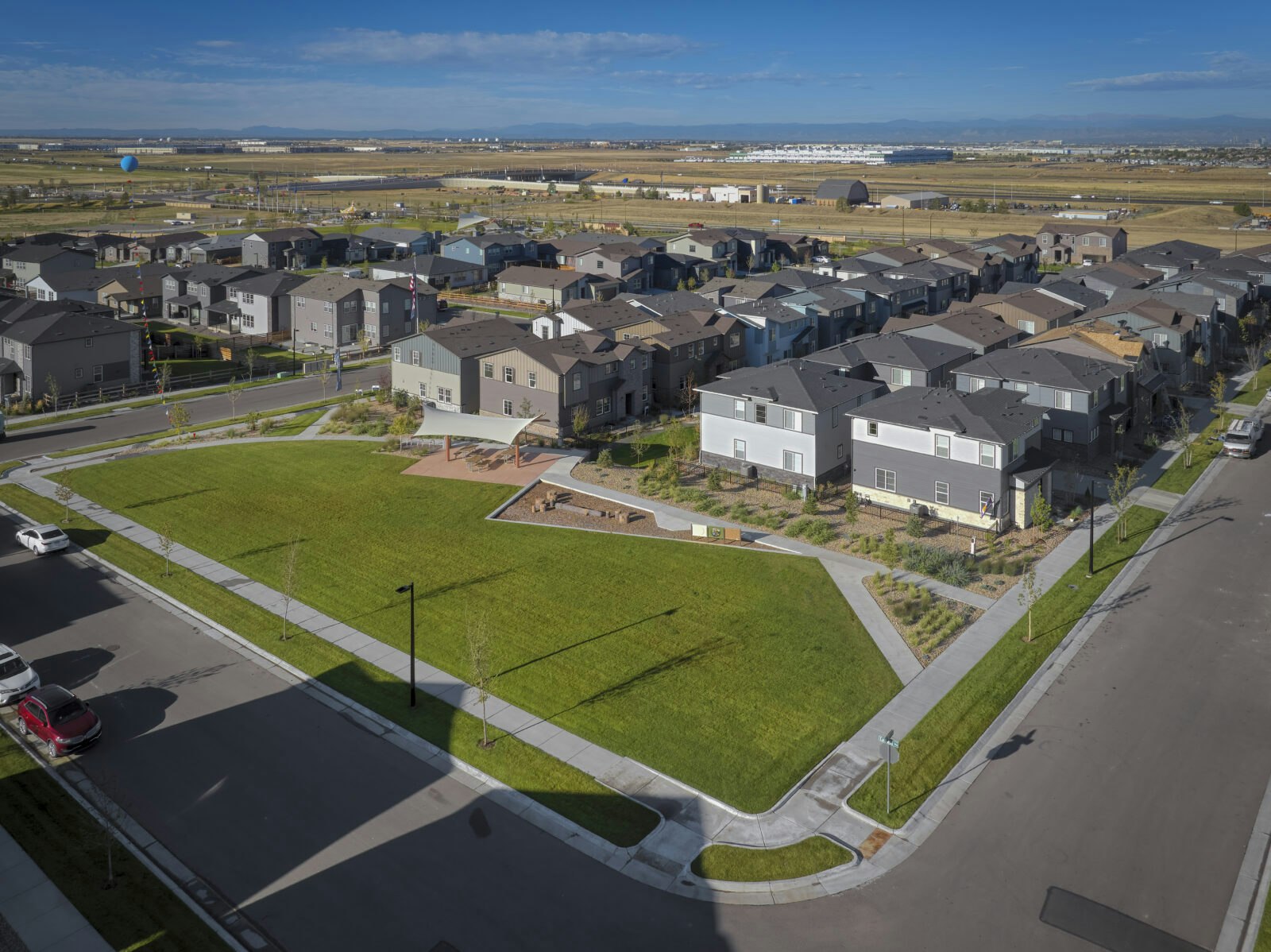A residential neighborhood with modern houses, green lawns, pathways, and roads. A balloon is visible in the distant sky.