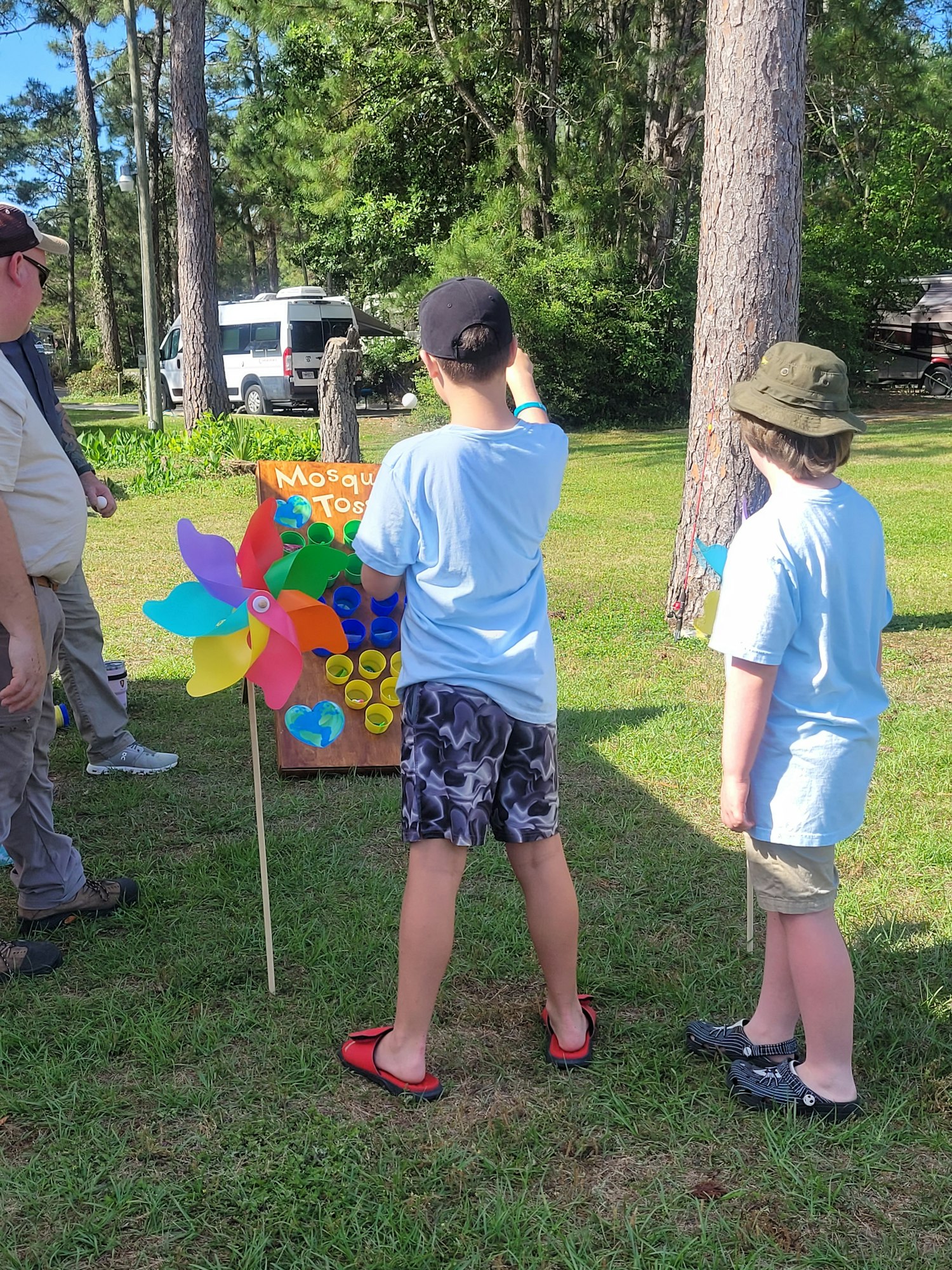 Two kids stand near a colorful pinwheel and a sign in a grassy area, possibly at an outdoor event or gathering.