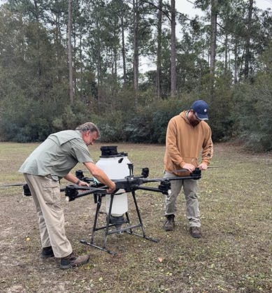 Two individuals are working together to assemble or adjust a large drone in a wooded outdoor setting.