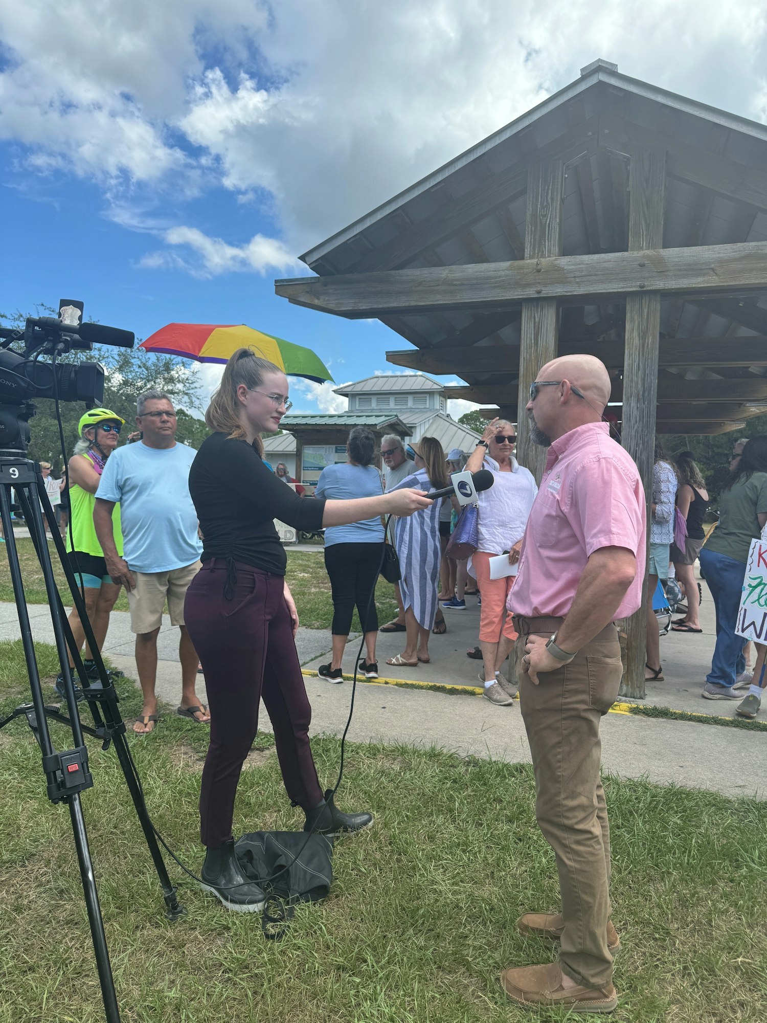 A news interview takes place outdoors with a reporter, a microphone, and a crowd in the background holding signs.