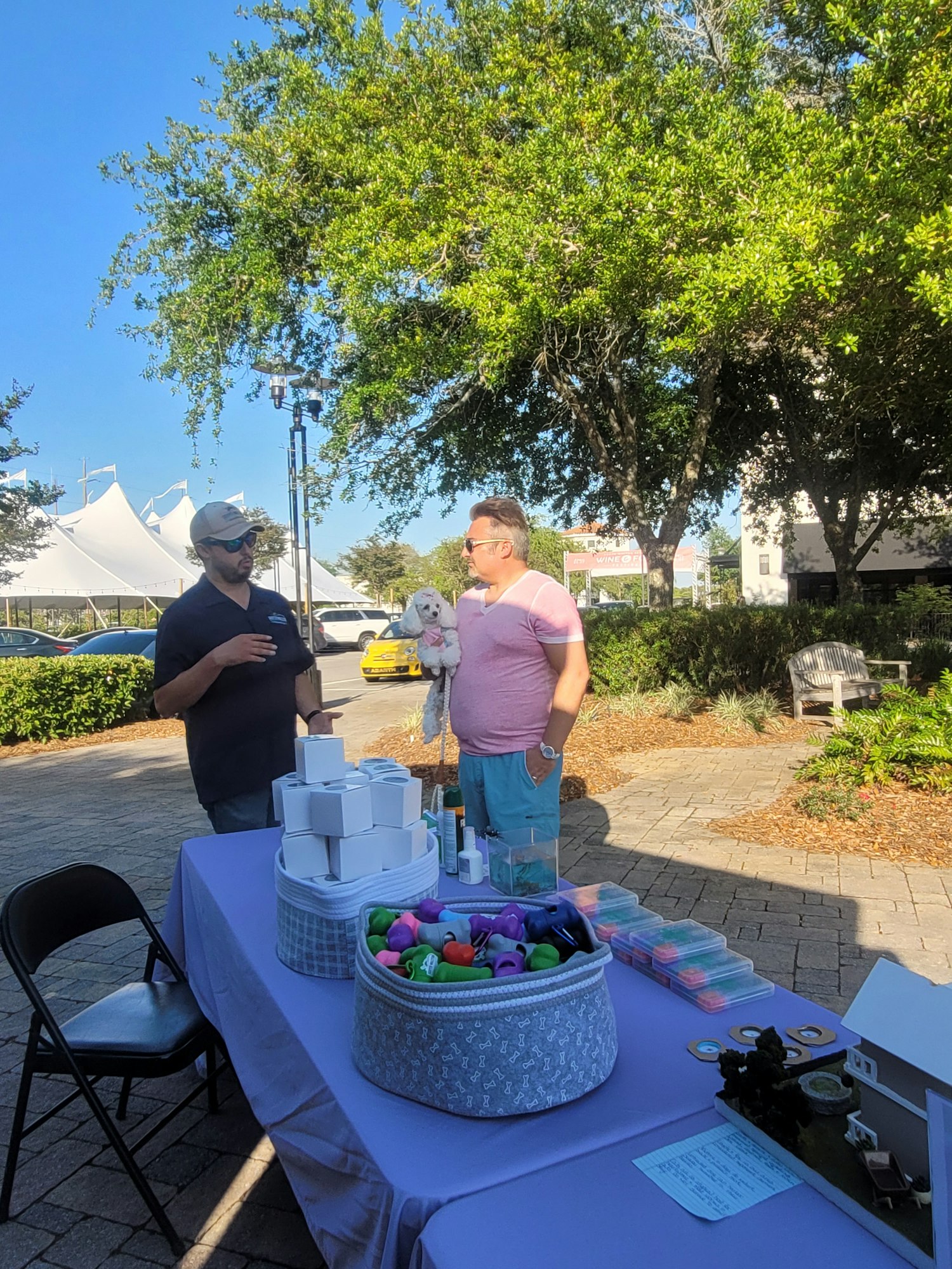 A market scene with two people chatting near a table filled with colorful items and a dog, under a sunny sky.
