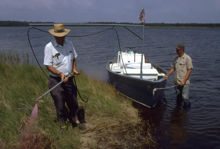 Two men work near a small boat on the water, one using a tool near the shore and the other adjusting the boat.