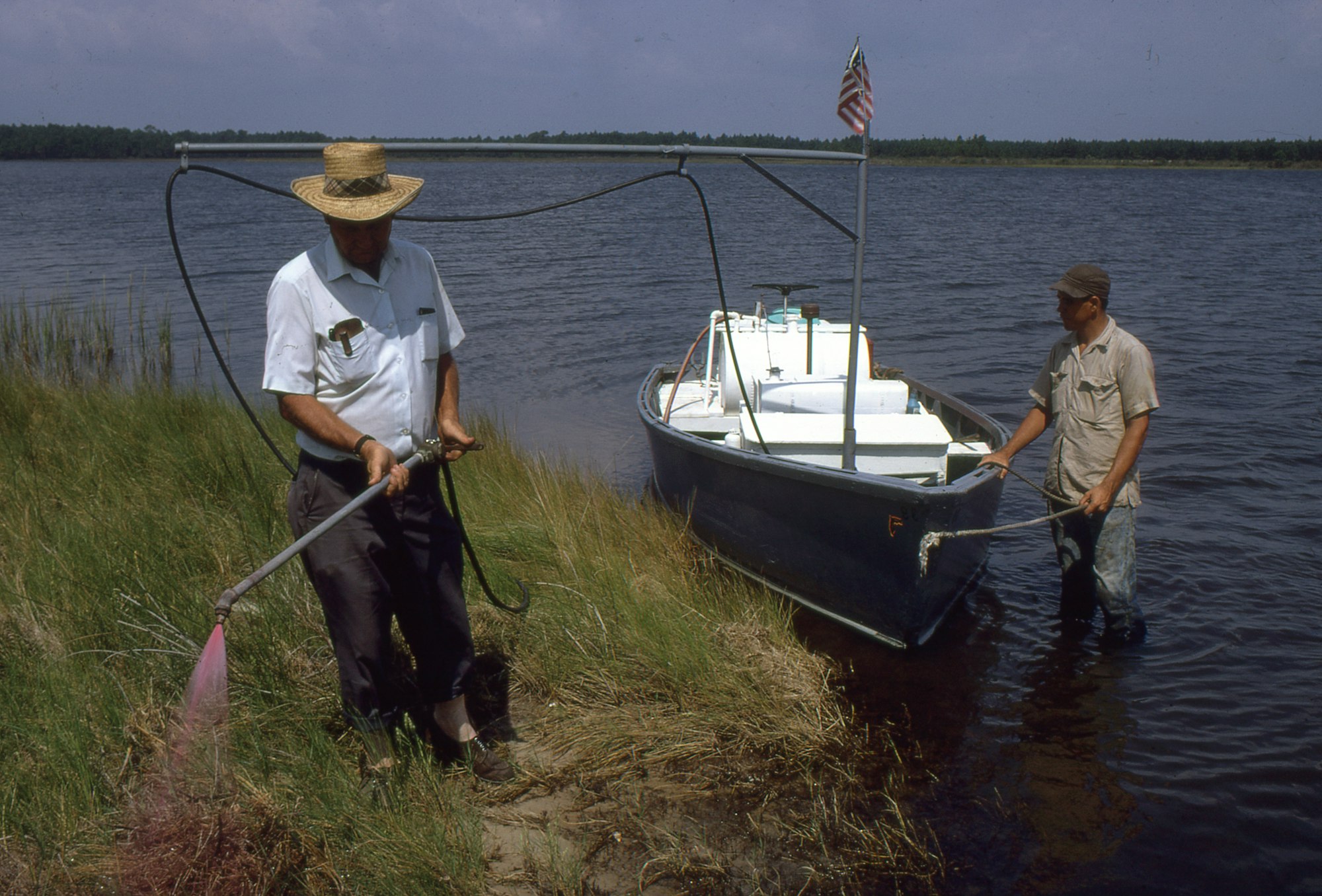 Two men work near a small boat on the water, one using a tool near the shore and the other adjusting the boat.