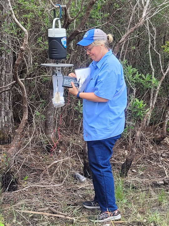 A person in a blue shirt and cap is setting up or working with equipment in a wooded area.