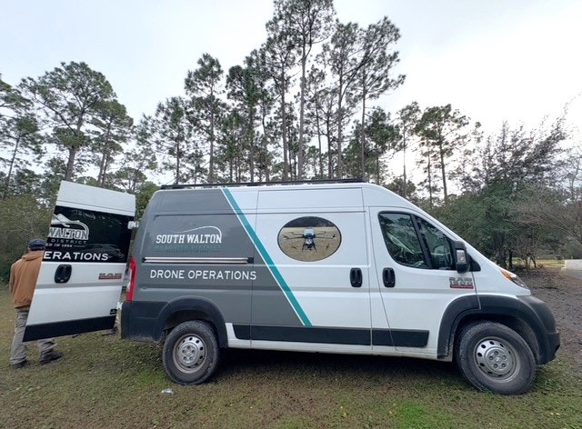 A van labeled "South Walton Drone Operations" parked in a forested area, with a person near the open door.