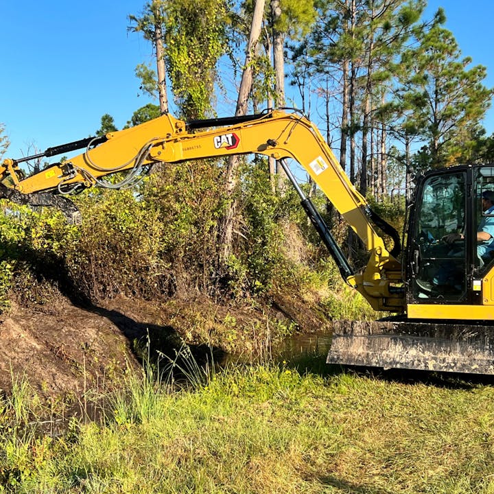 A yellow excavator is working near a waterway, clearing vegetation, with a person operating it inside.