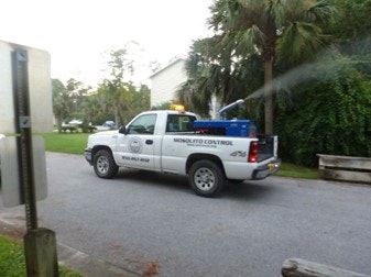 A white pickup truck marked "Mosquito Control" sprays insecticide on a street lined with trees and greenery.