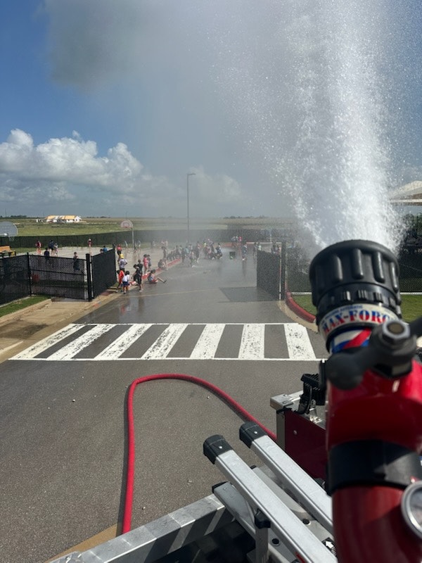 A fire truck hose spraying water onto a group of children playing on a sunny day in a fenced area.