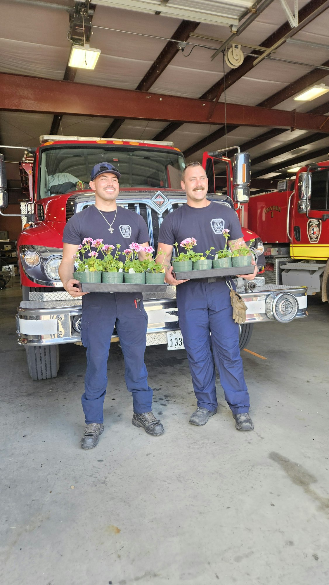 Two people in uniforms hold trays of small plants in front of a fire truck inside a garage.