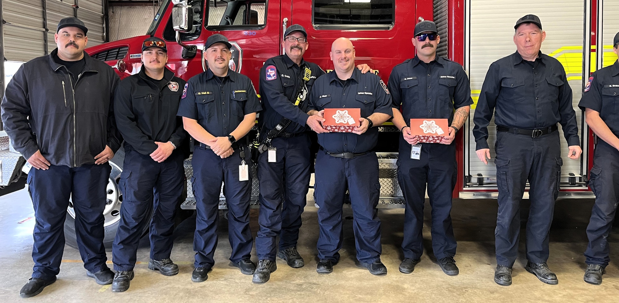 A group of firefighters poses together in front of a fire truck, holding gift boxes and smiling.