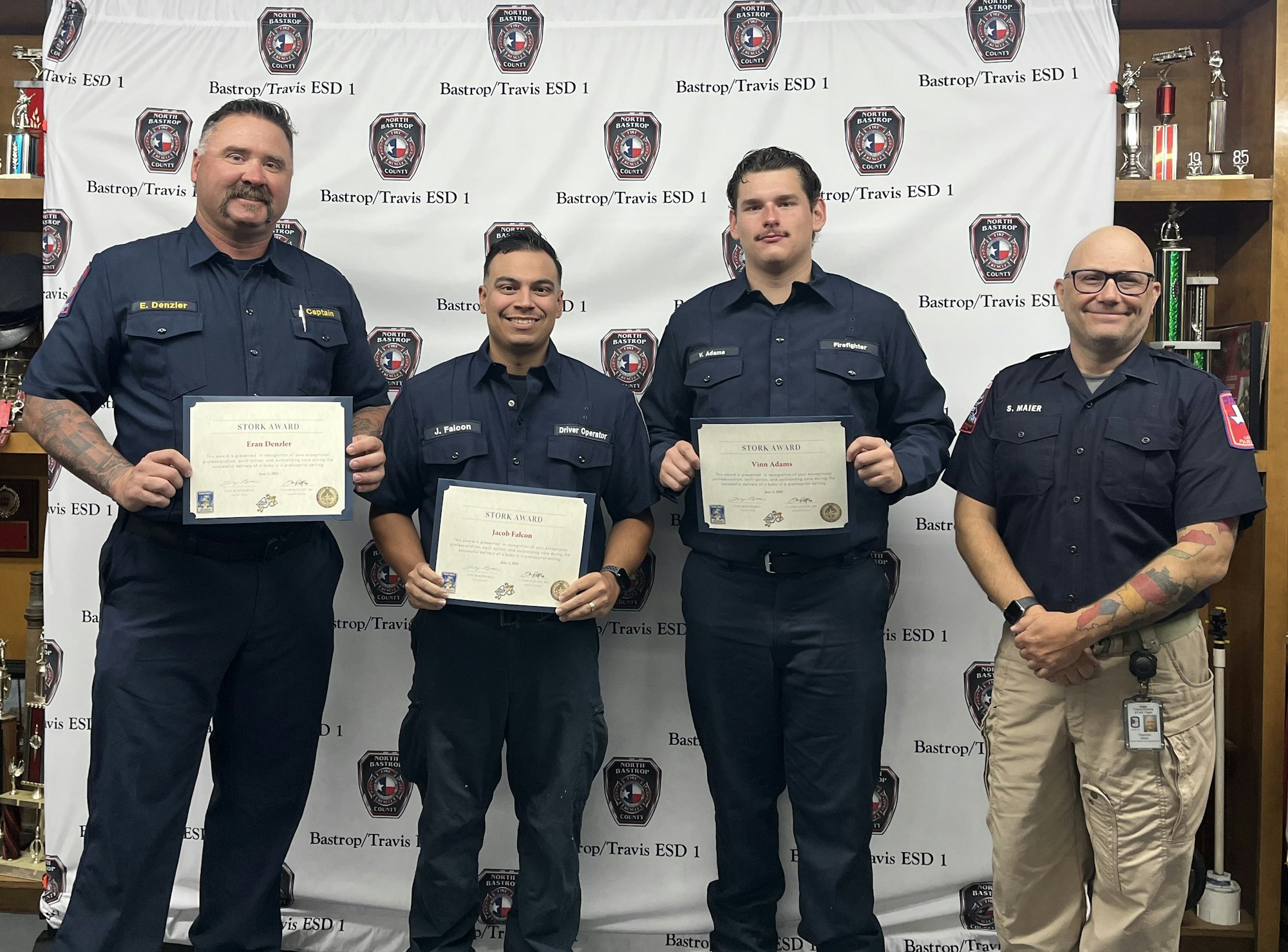 Four people in uniform, three holding certificates, standing in front of a "Bastrop/Travis ESD 1" backdrop.