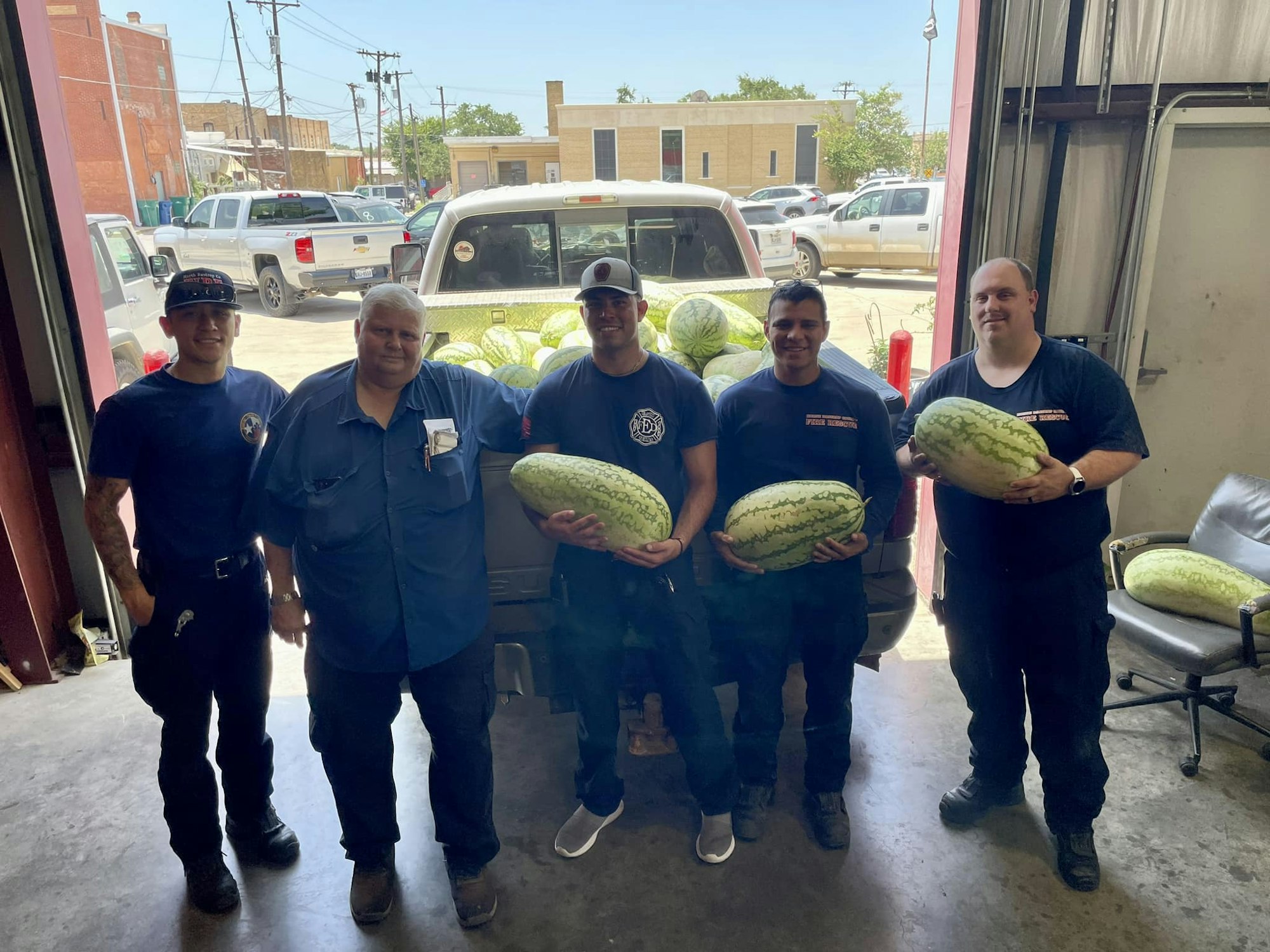 Five people, likely firefighters, holding watermelons in a garage with a truck in the background.