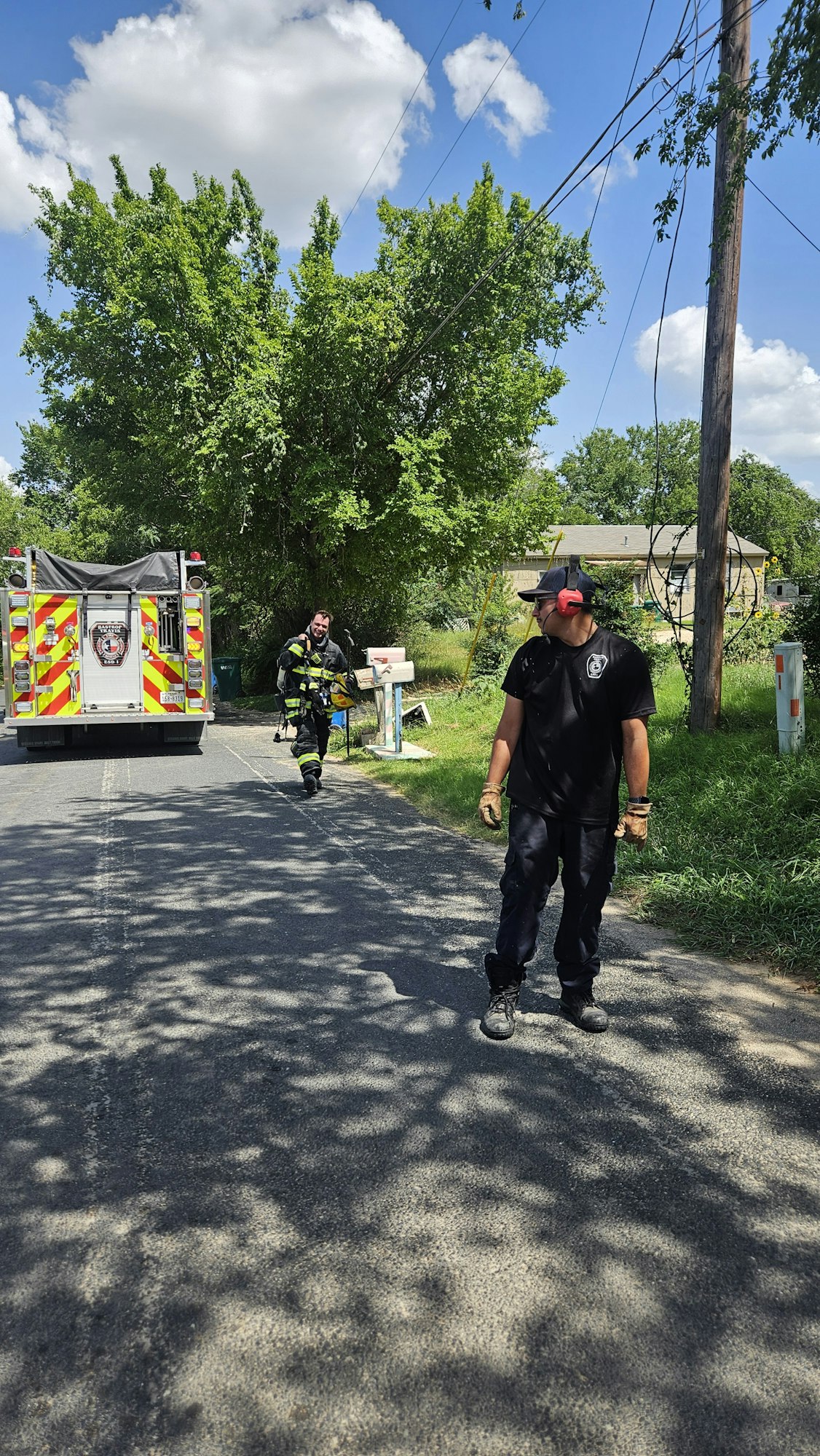 Two people near a utility truck, one in black clothes with a red headset, the other in safety gear, are on a shaded street with trees.
