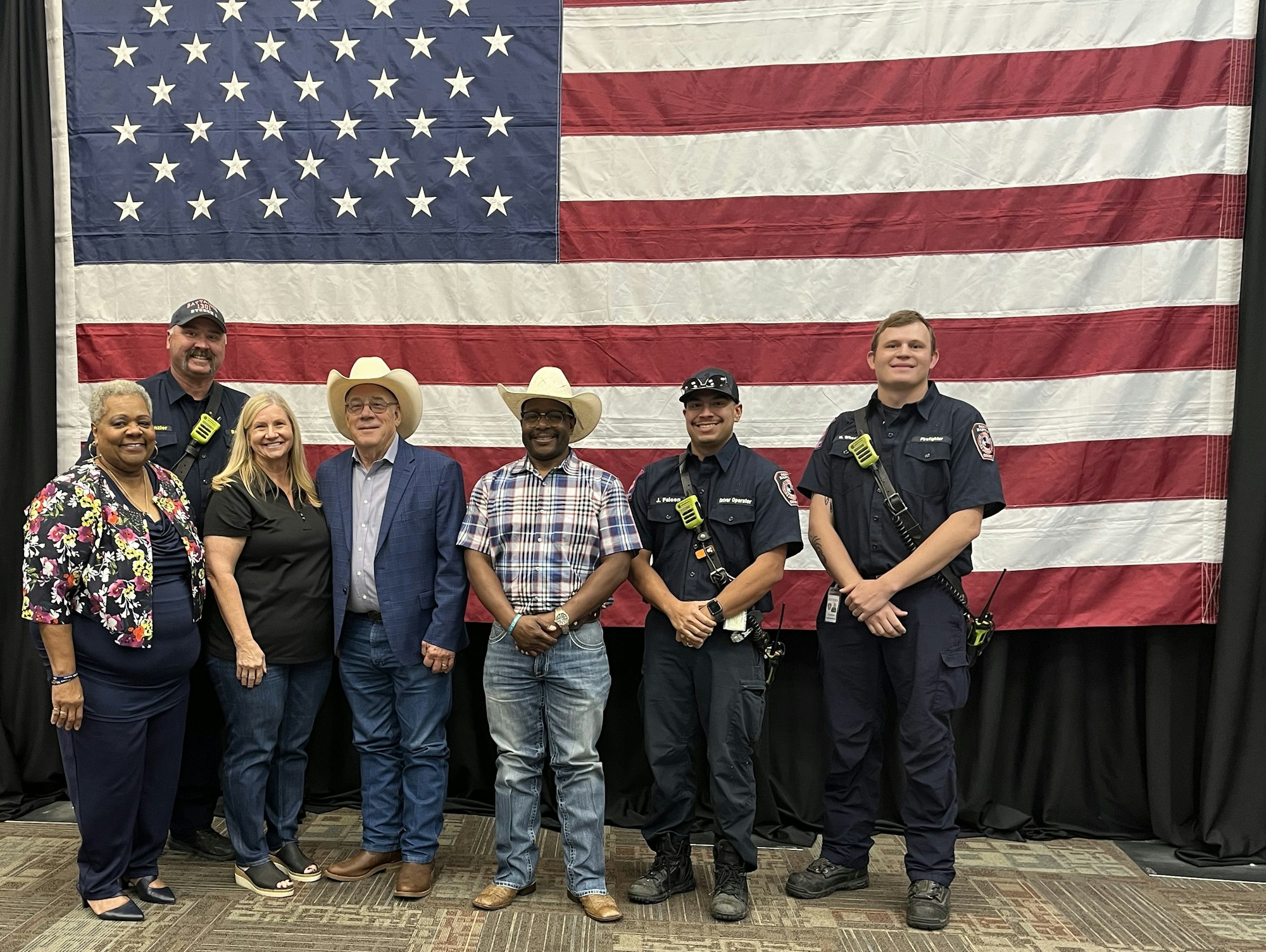 A group of individuals, including first responders and community members, poses in front of an American flag backdrop.