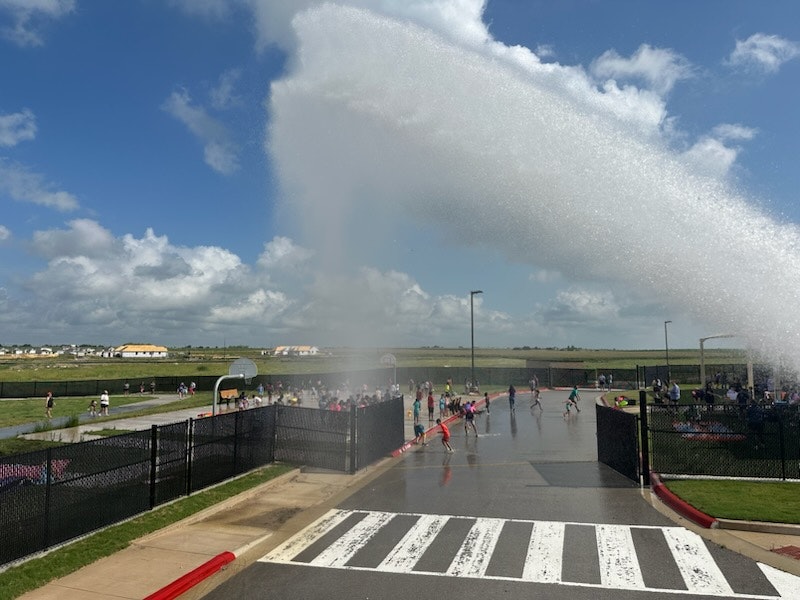 People enjoying water spray on a playground under a blue sky.