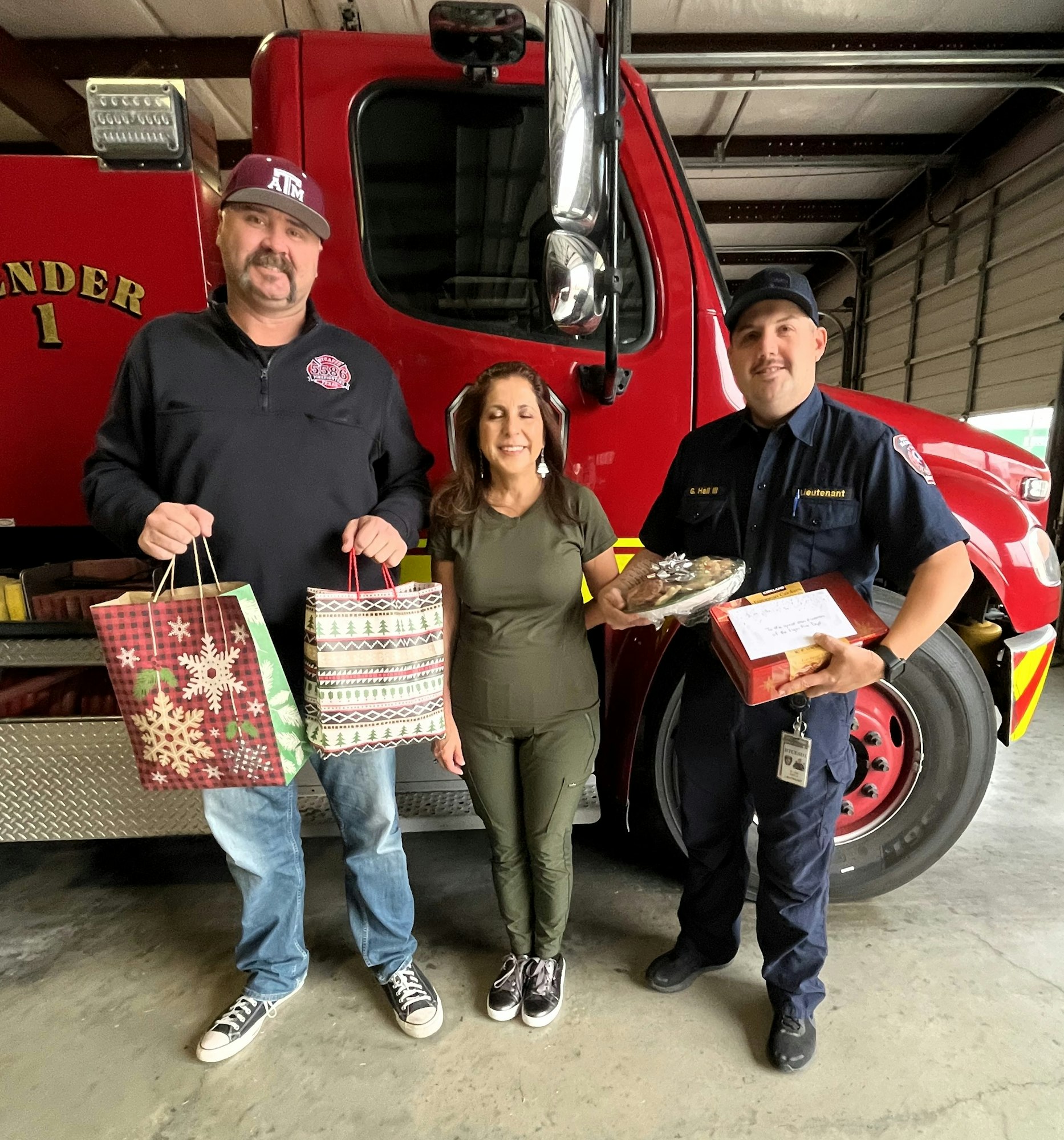 Three people are standing in front of a fire truck, holding gifts and food, smiling together.