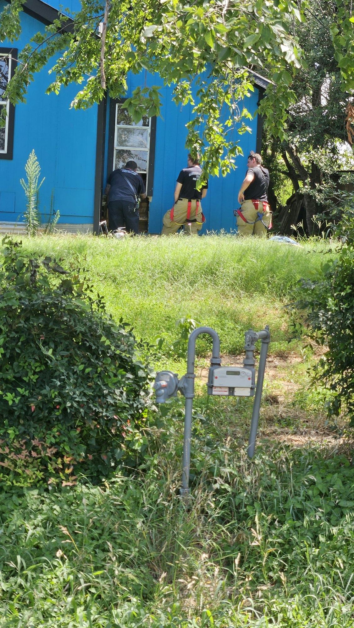 Two people, possibly utility workers, are beside a blue building surrounded by greenery. There's a gas meter in the foreground.