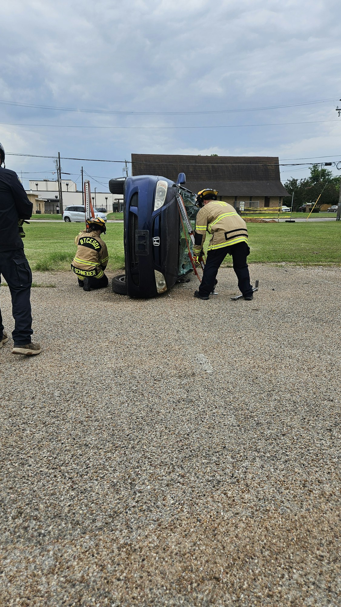 Firefighters working near an overturned blue car on a gravel surface.