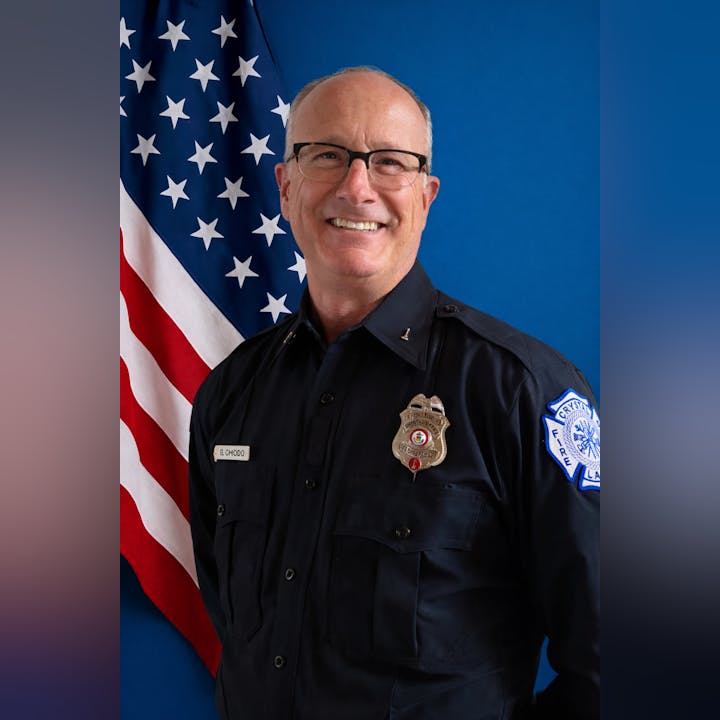 A smiling person in a fire department uniform stands in front of an American flag backdrop.