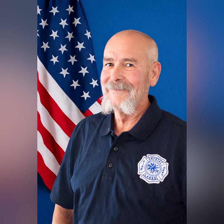 A smiling older man in a navy polo with a fire department badge, standing in front of an American flag.