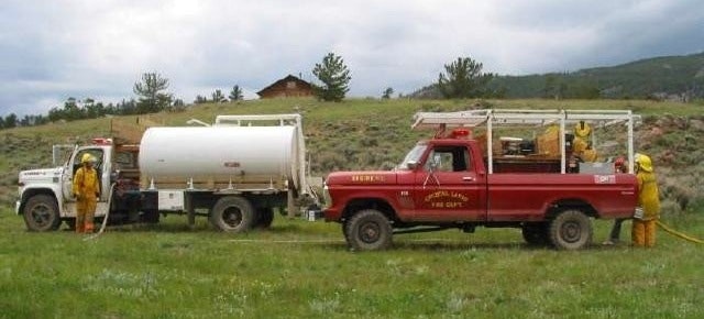 Two fire trucks in a grassy area, one red and one white, with firefighters in yellow suits handling hoses.
