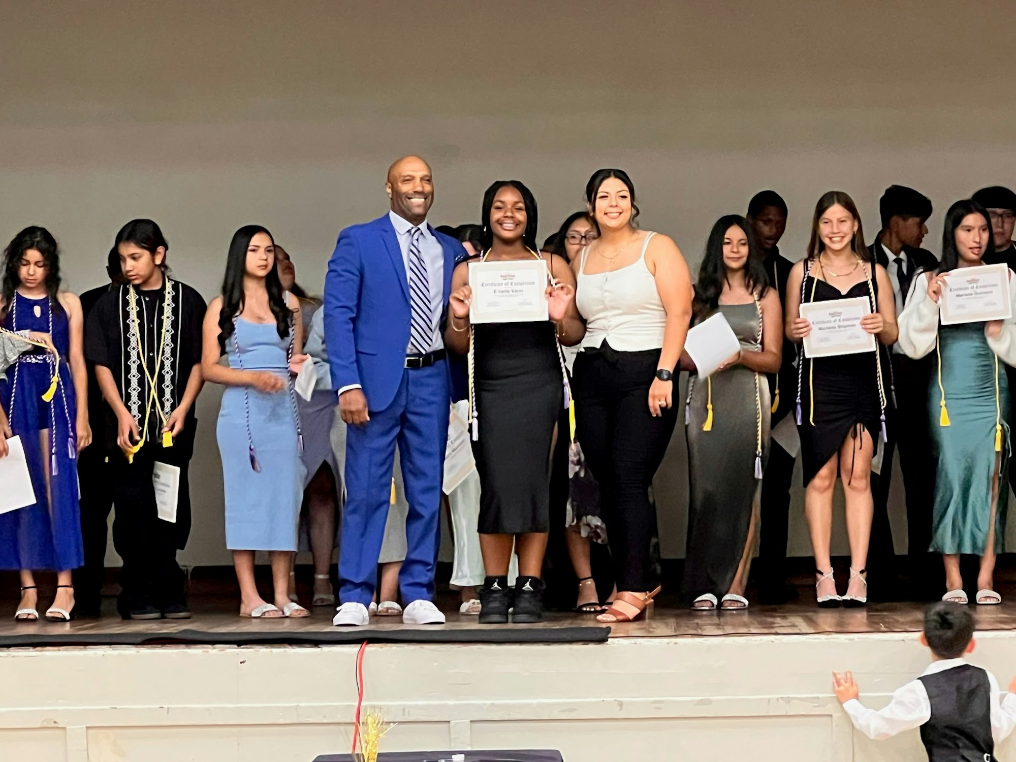 A group of students with certificates on stage, celebrating an academic achievement, with an adult overseeing the event.