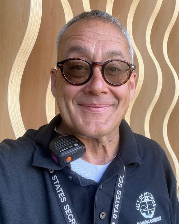 A person smiling, wearing round glasses and a "City of Pittsburg Planning Commission" shirt, with a radio on the collar.