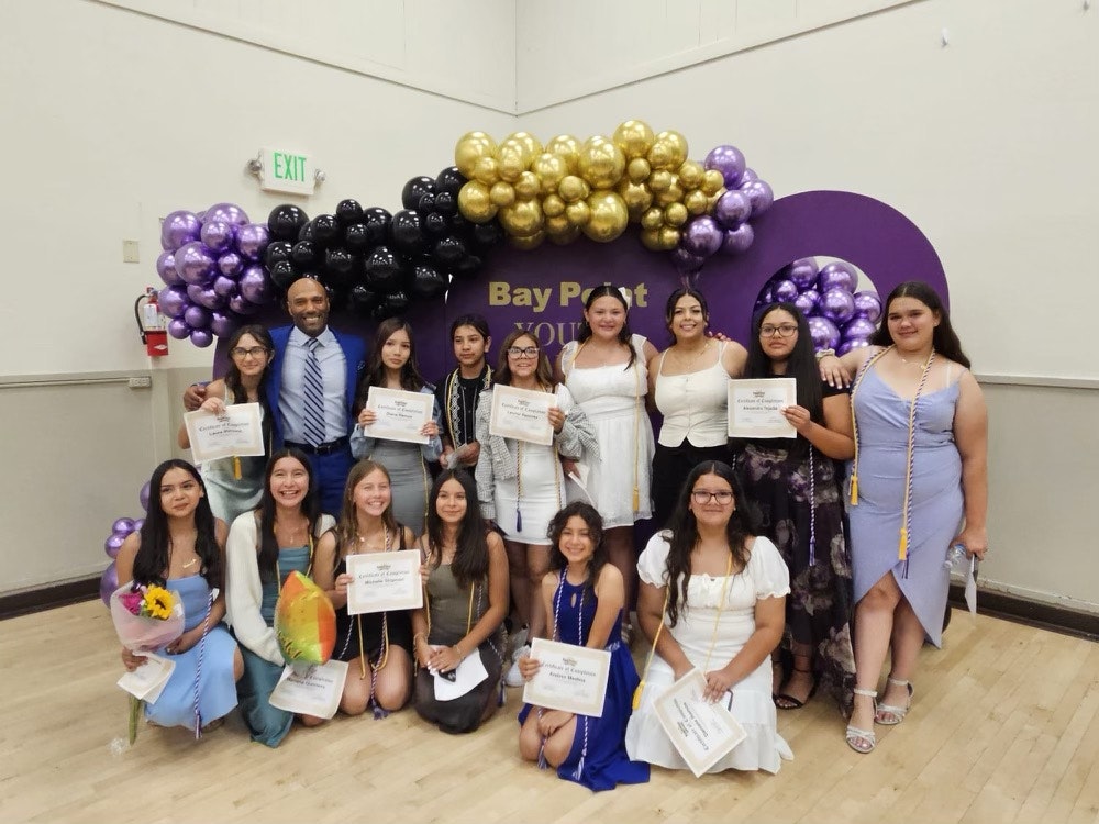 A group of people celebrating an achievement with certificates, balloons, and decorative colors in the background.