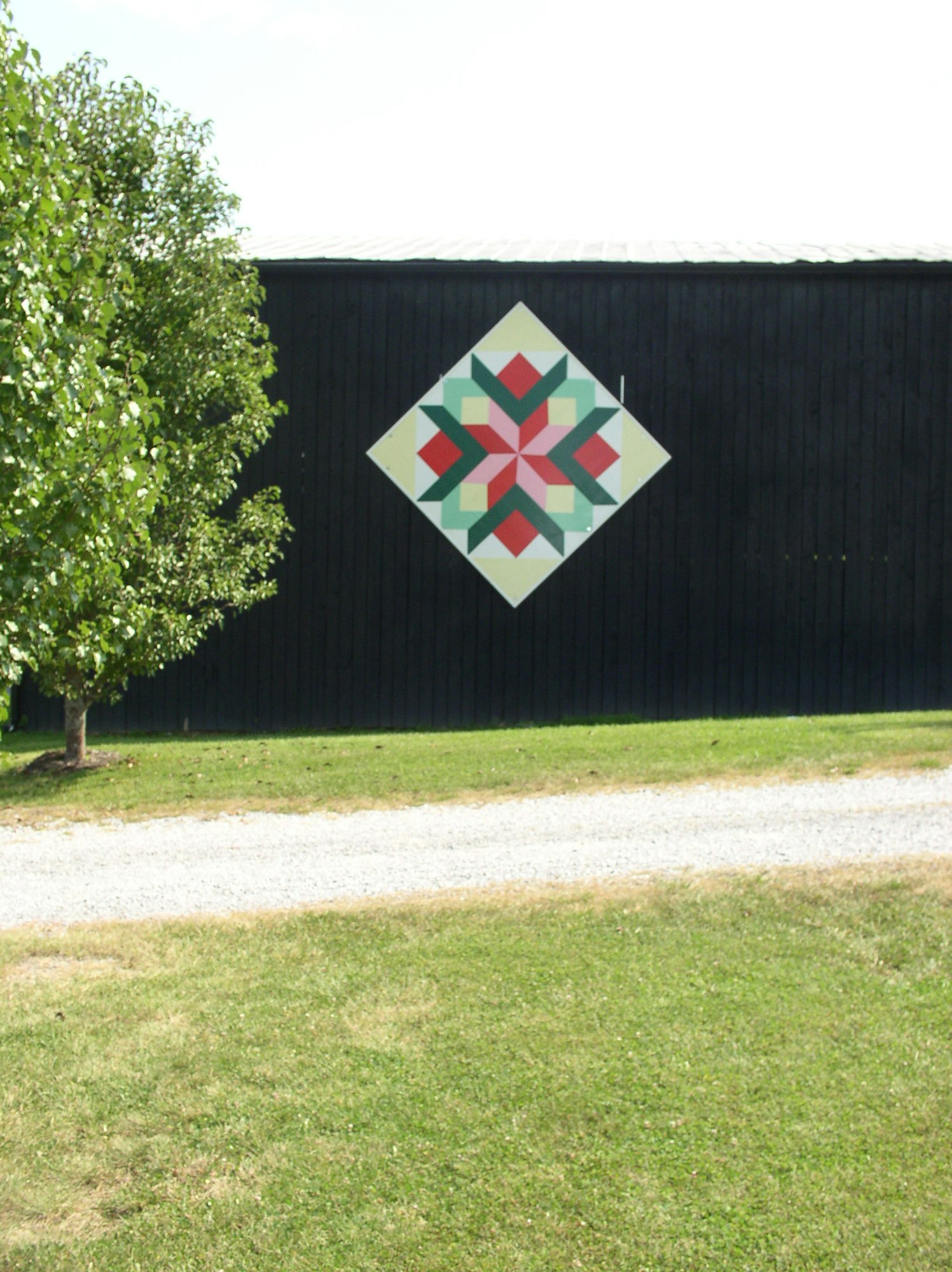 A barn quilt with a geometric pattern on a black wall, grass, a tree, and a gravel path in front.
