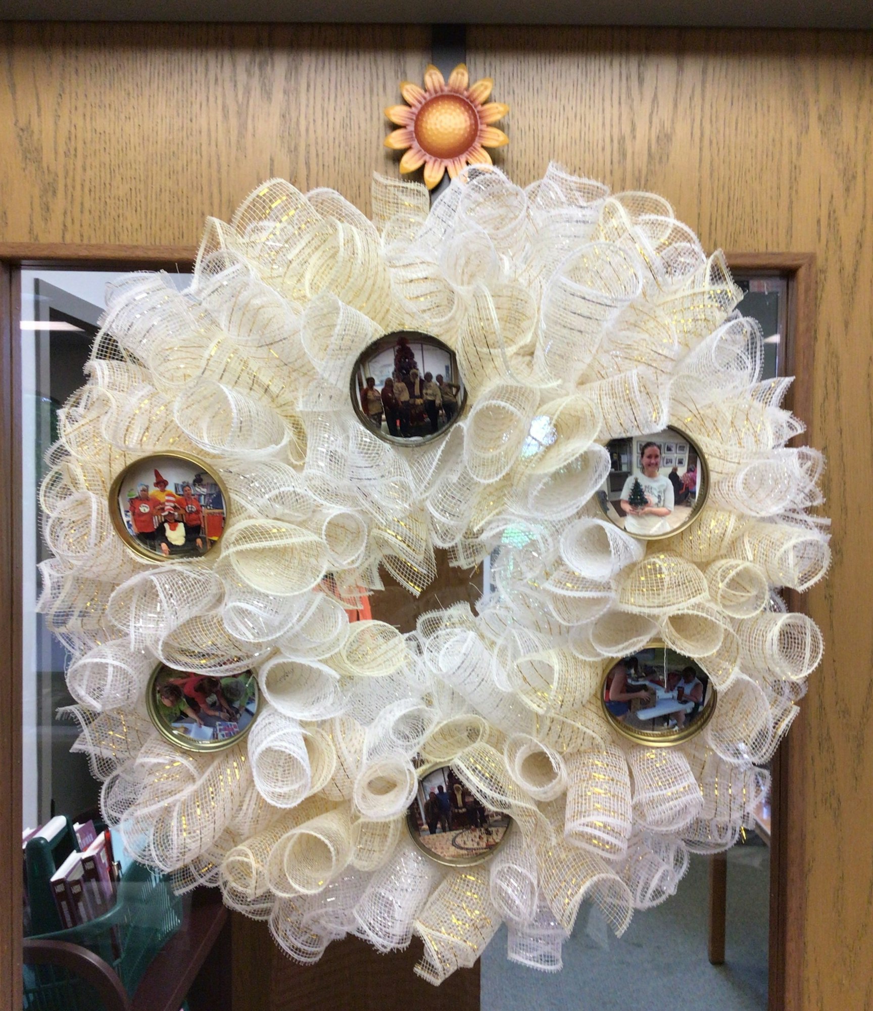 A decorative white fabric wreath featuring circular frames with photos, topped with a sunflower, hanging on a door.