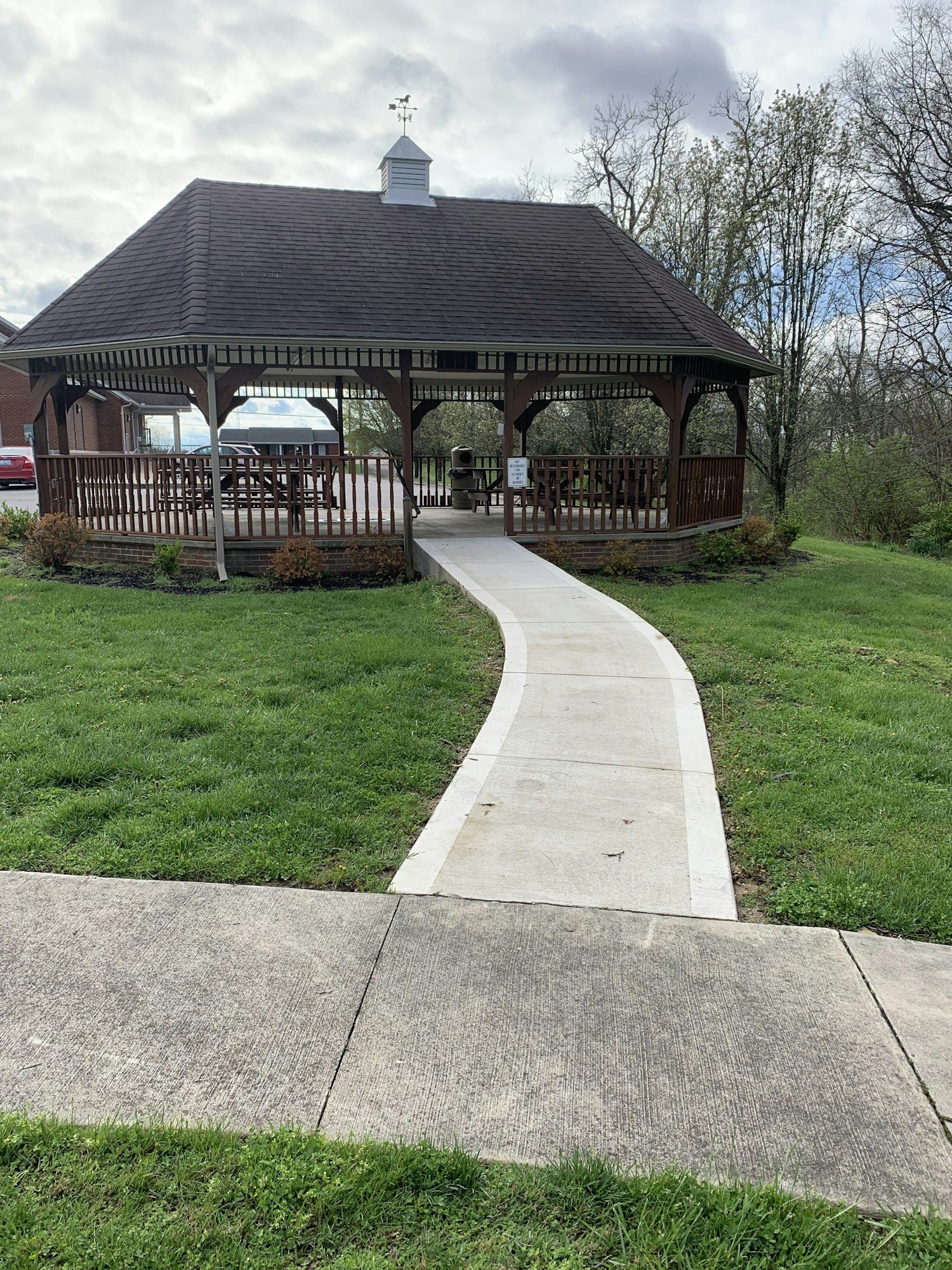 A gazebo with a path leading to it, surrounded by grass and trees under a cloudy sky.