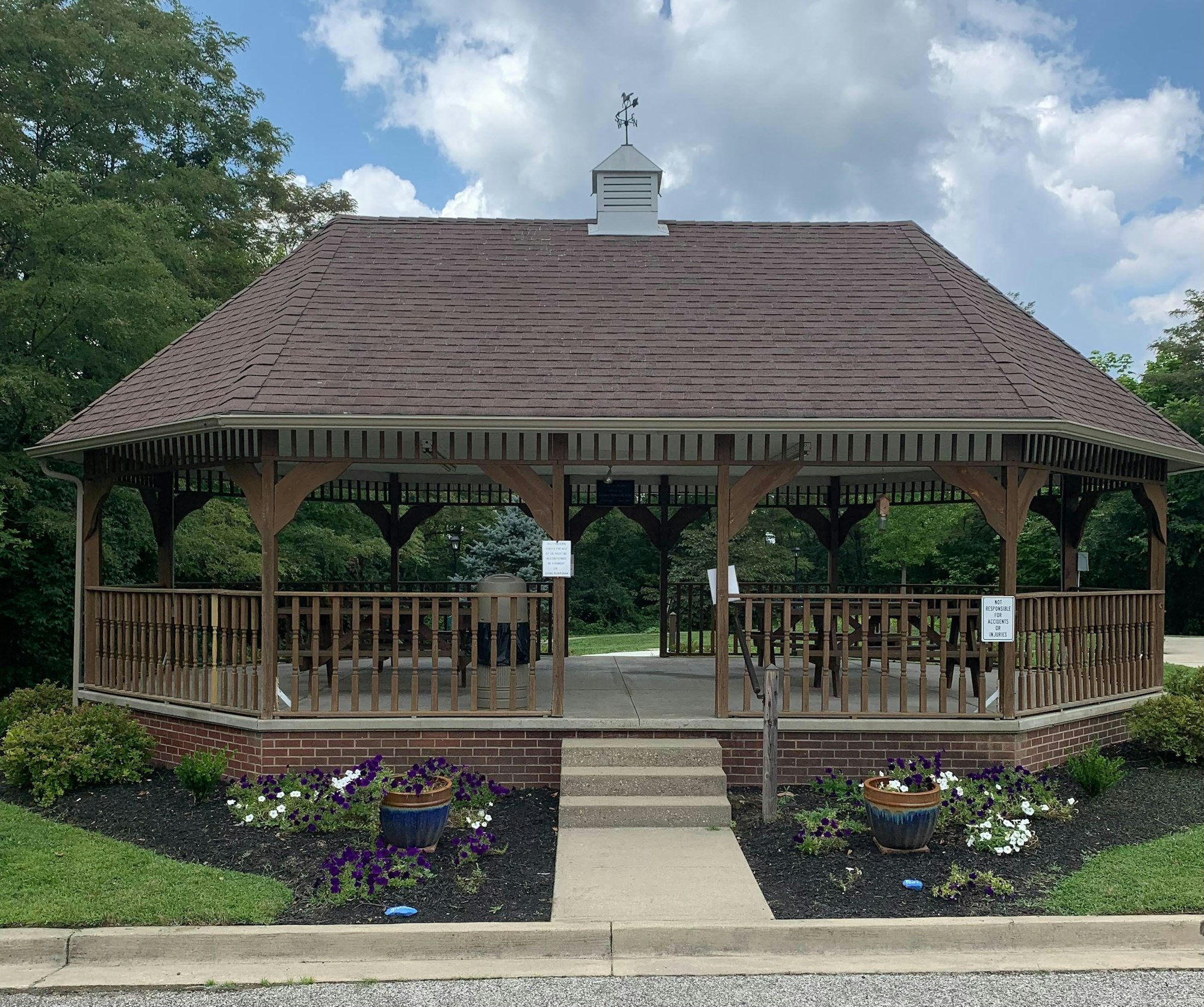 A wooden gazebo with a shingled roof, surrounded by plants and flowers, set against a background of trees and a partly cloudy sky.