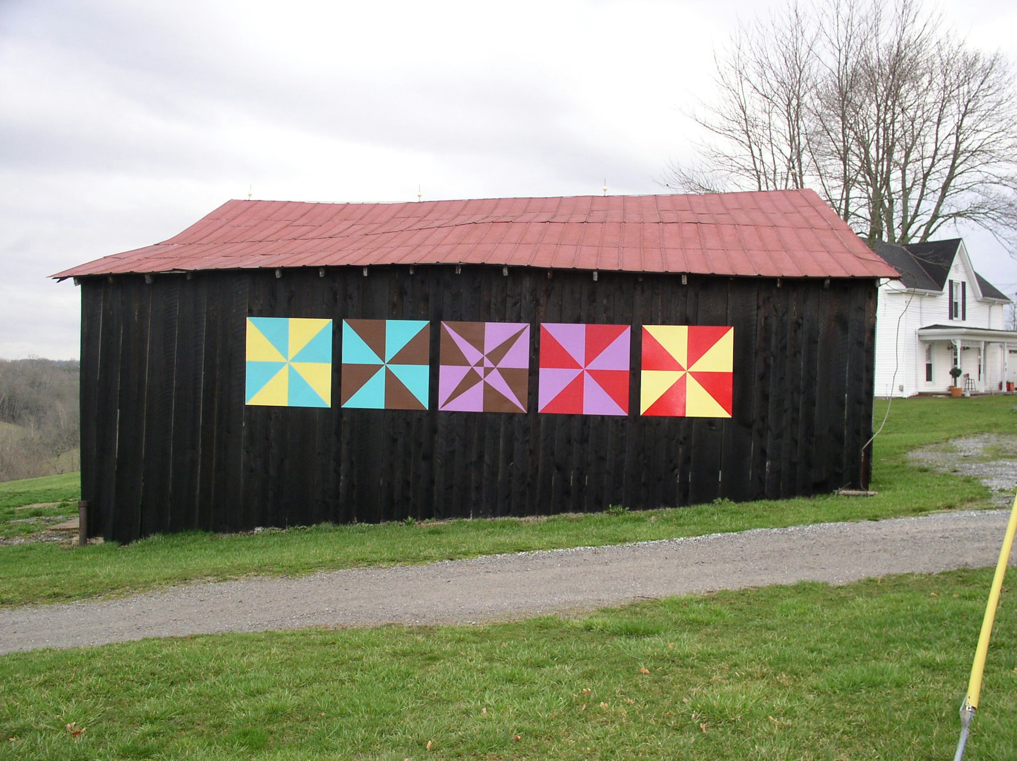 A barn with a red roof and six colorful quilt patterns on its black wall, situated in a grassy landscape.