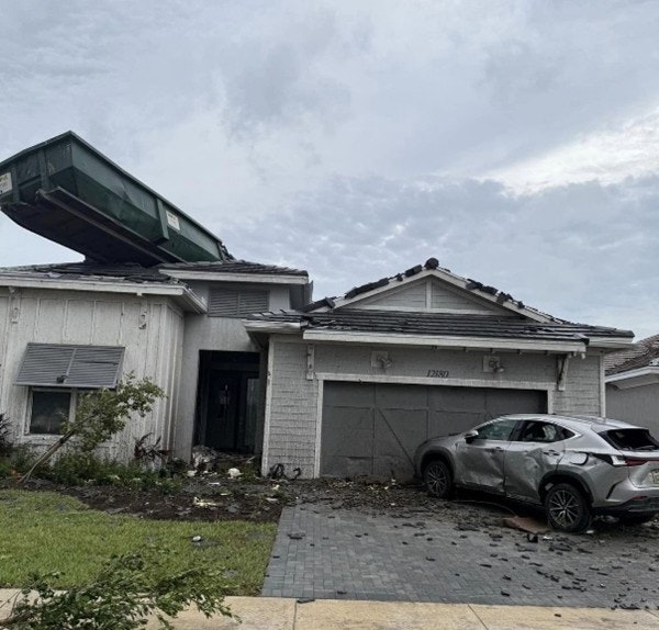 A damaged house with a car on the driveway and a large container on the roof.