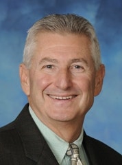 A smiling man with grey hair in a suit and tie against a blue background.