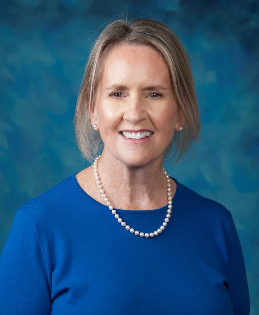 A smiling woman with pearl necklace, wearing a blue top, against a blue textured background.