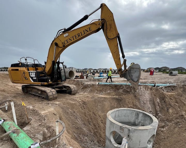 Excavator on a construction site with workers, trenches, and concrete pipes.