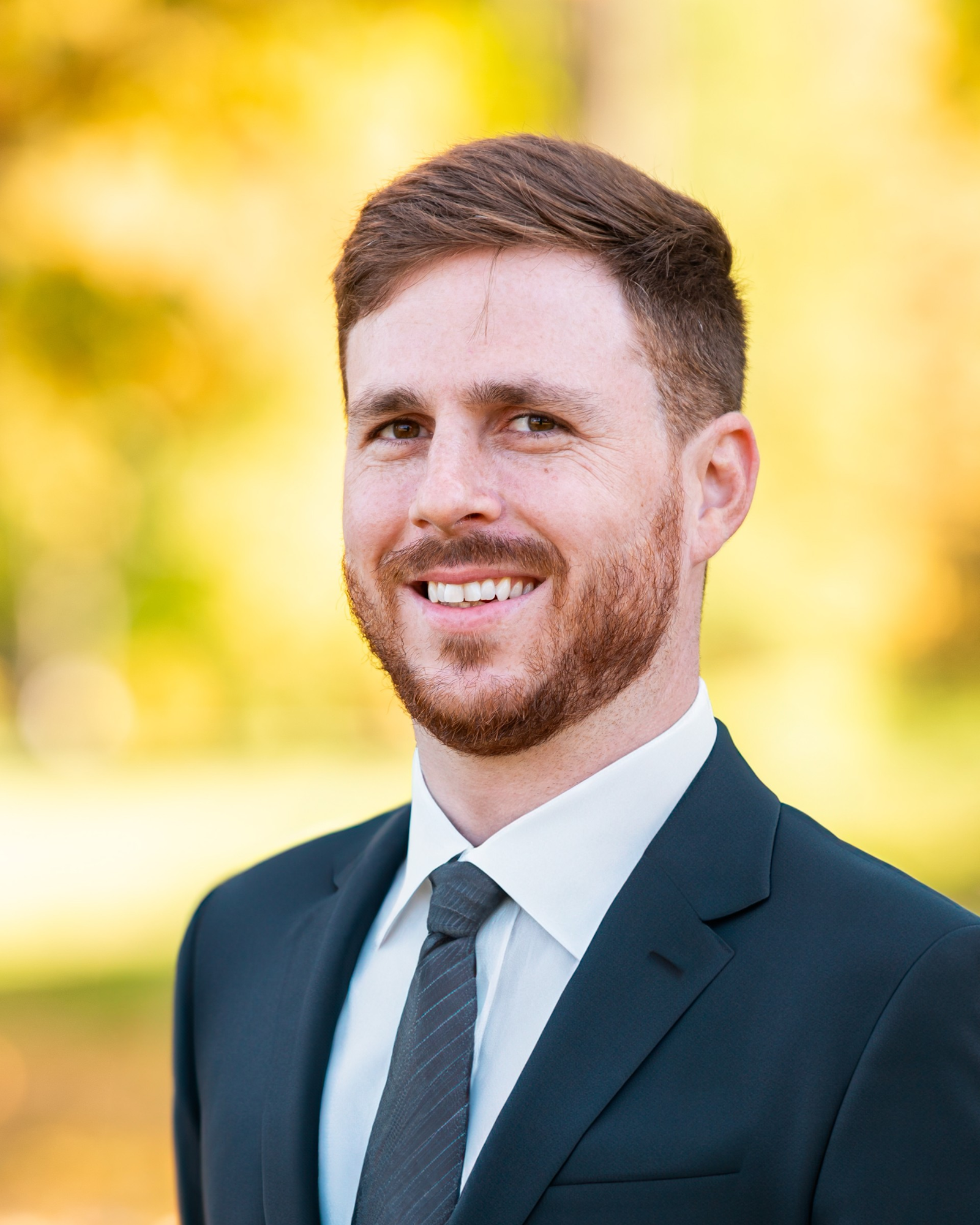A man in a suit, smiling, with a blurred natural background.
