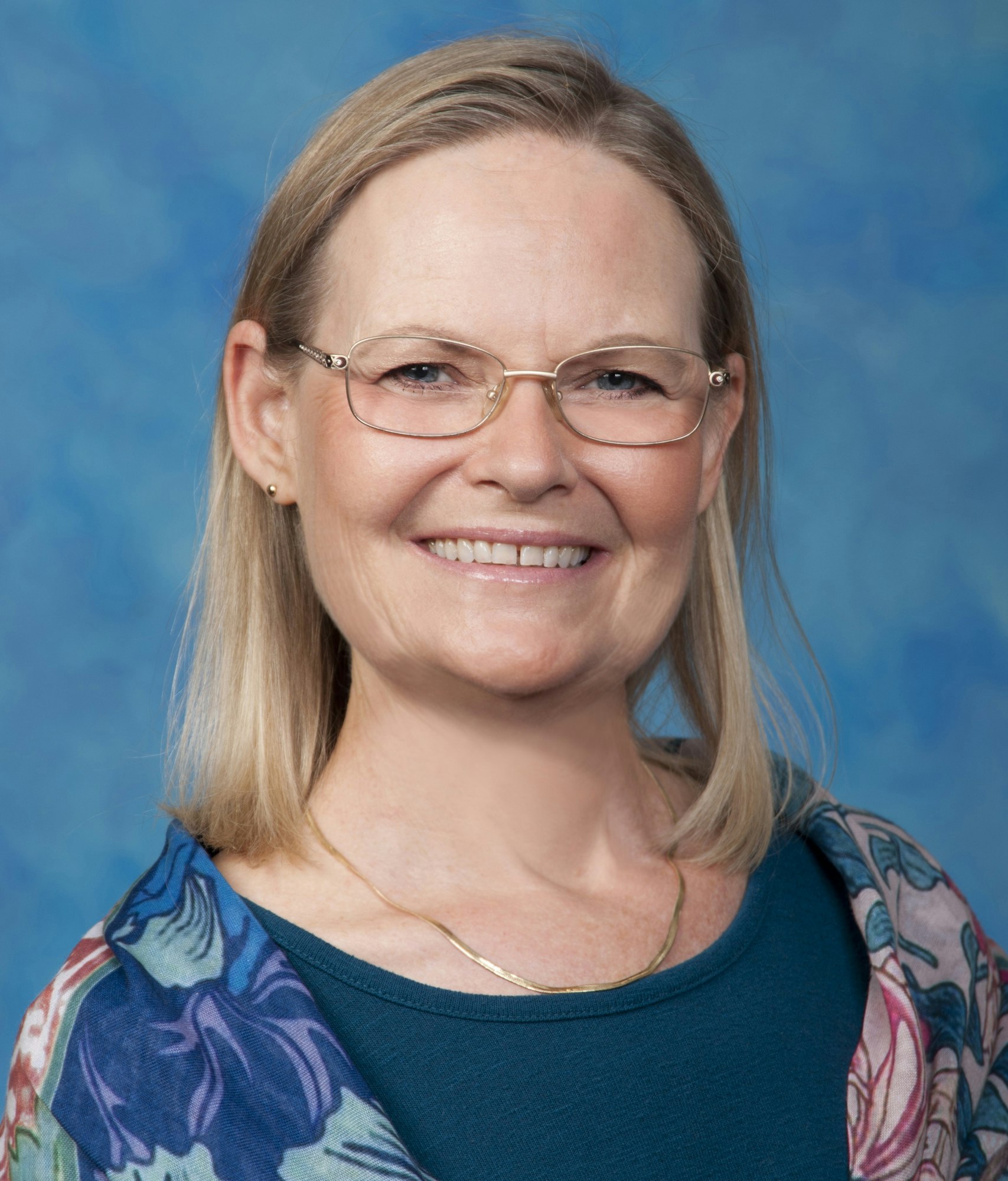 Portrait of a smiling woman with glasses, wearing a floral outfit against a blue background.