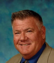 A smiling man with a blue shirt, suit jacket, and tie against a mottled blue backdrop.