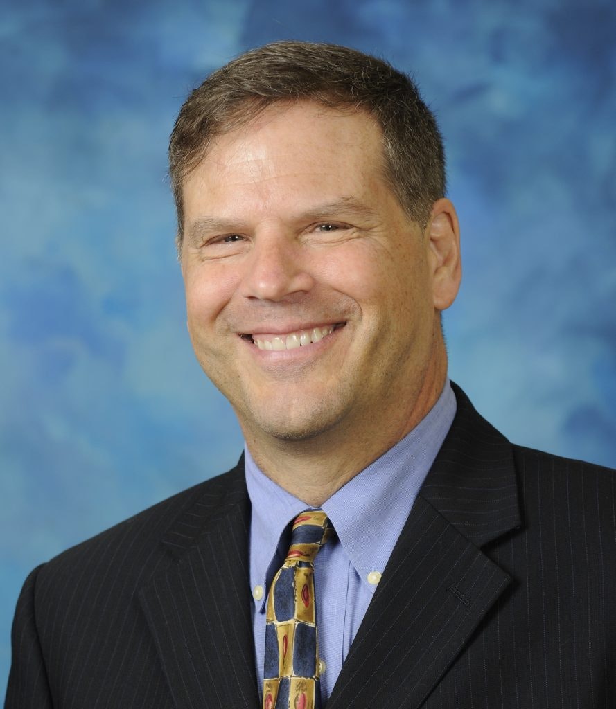 A smiling person with a blue patterned background, wearing a suit and a tie.
