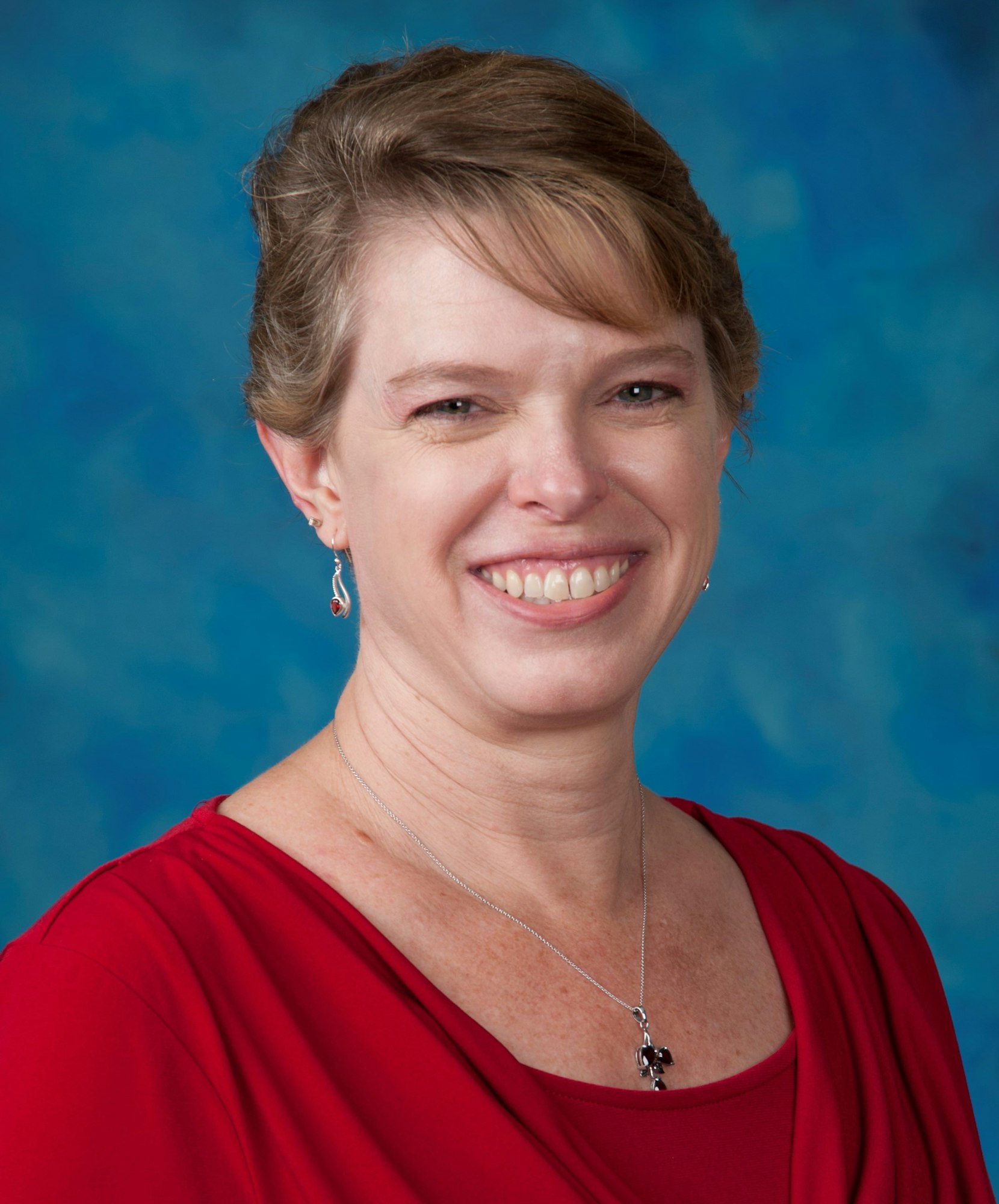 A smiling woman with short hair, wearing a red top, earrings, and a necklace against a blue backdrop.