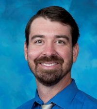 A portrait of a smiling man with a beard, wearing a blue shirt and patterned tie against a blue background.