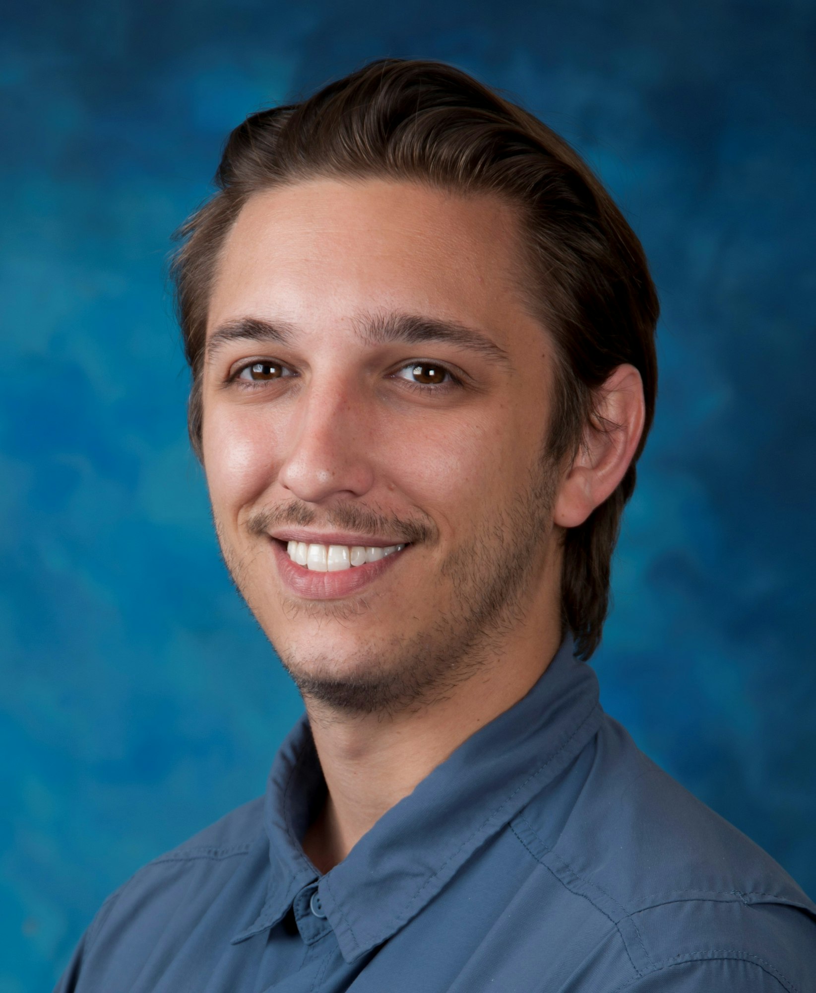 A smiling man with a goatee and a blue shirt against a blue background.