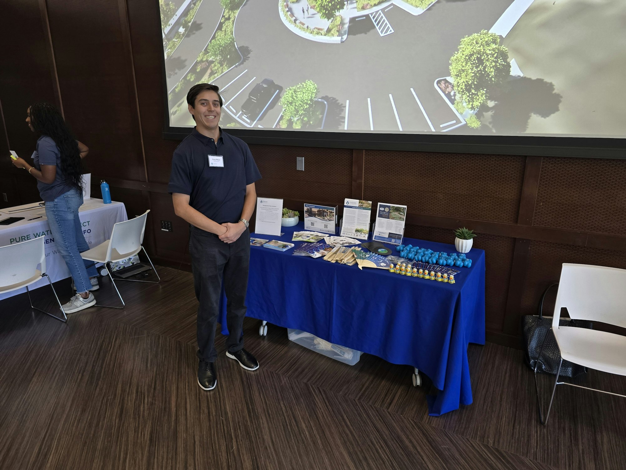 A man stands in front of a table with informational materials and giveaways, while a woman is busy in the background.