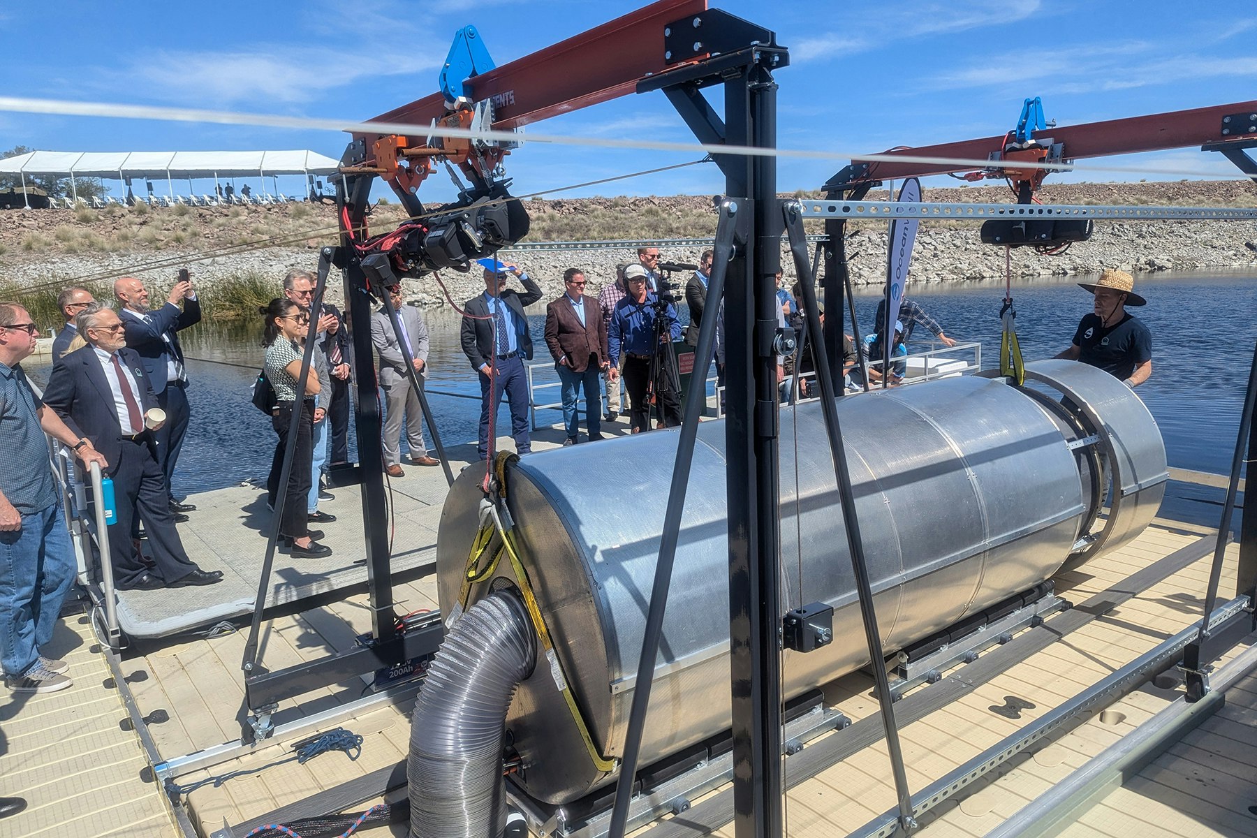 A group of people observes machinery on a waterfront platform, featuring a large metal cylindrical structure.